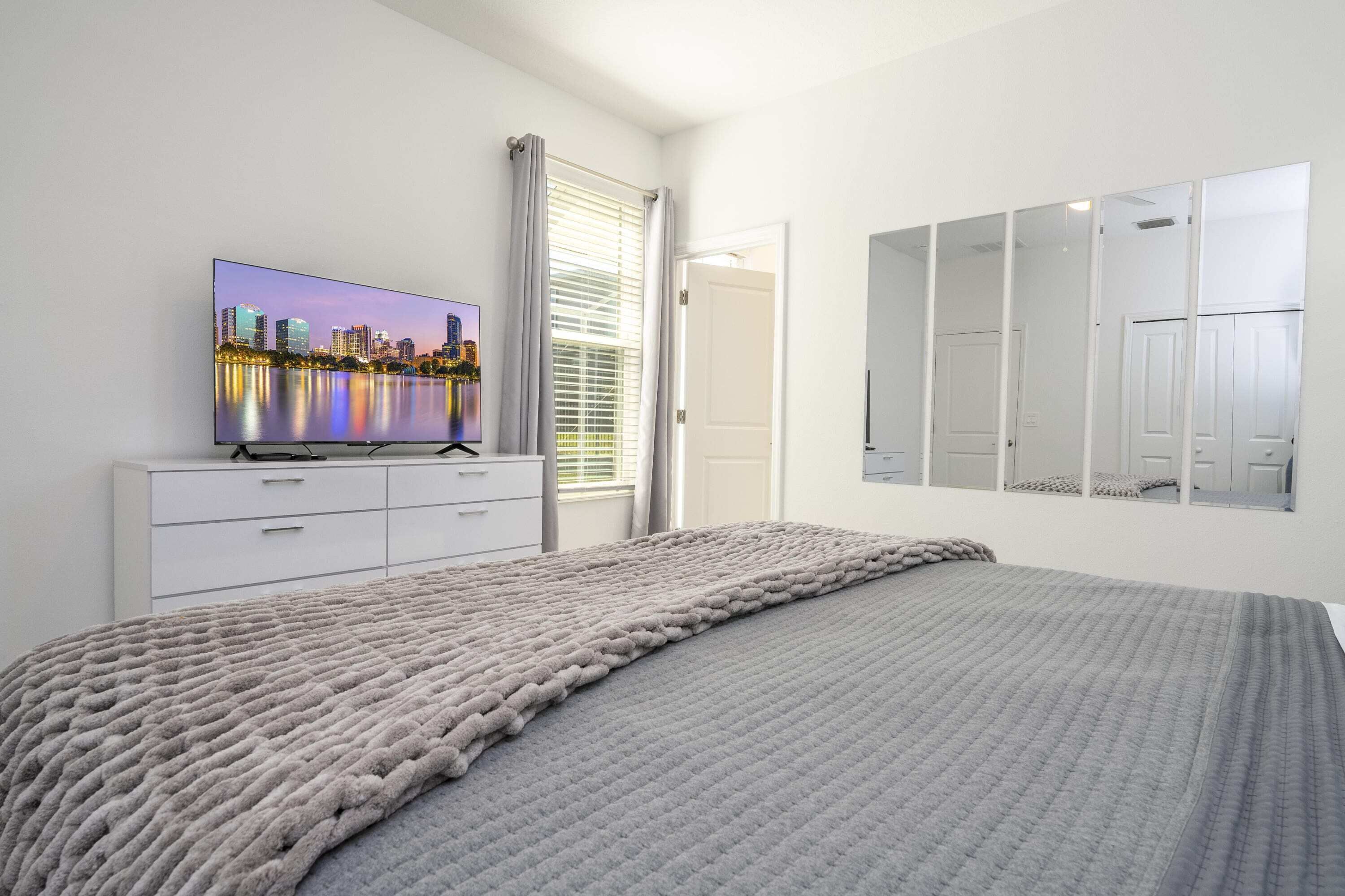 Een slaapkamer met een bed, een televisie op een dressoir, een raam met gordijnen en een spiegel aan de muur.