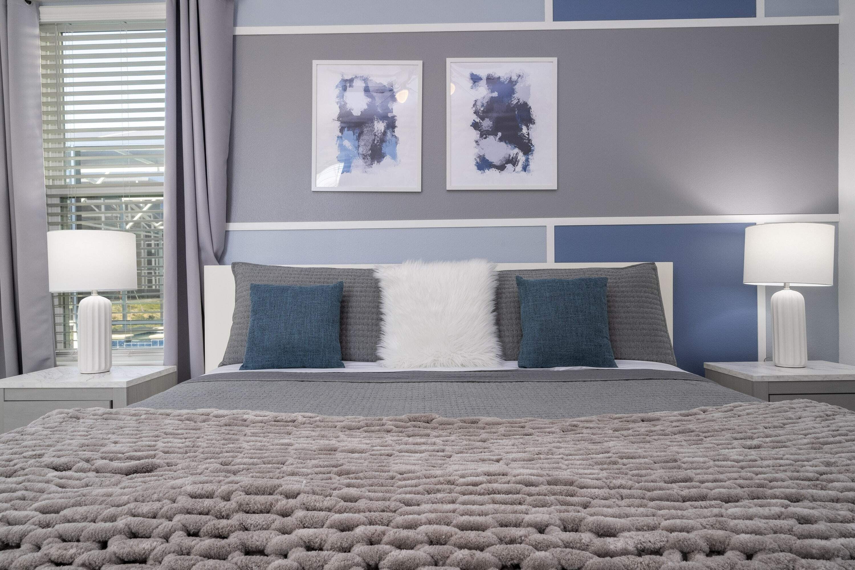 A bedroom with a large bed, two nightstands, a window with curtains, and abstract art on the wall.