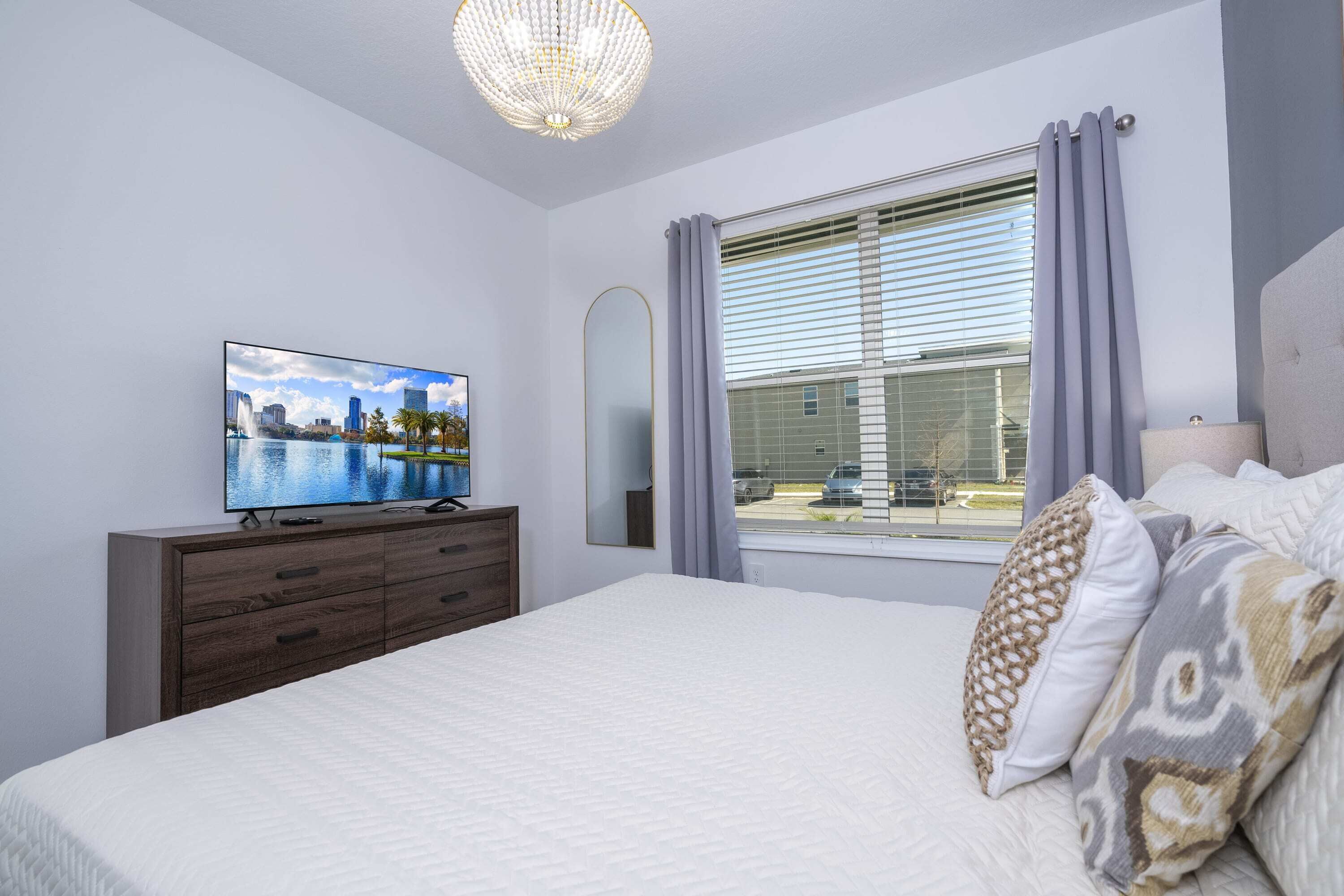 A bedroom with a bed, a television on a dresser, a window with curtains, and a chandelier.