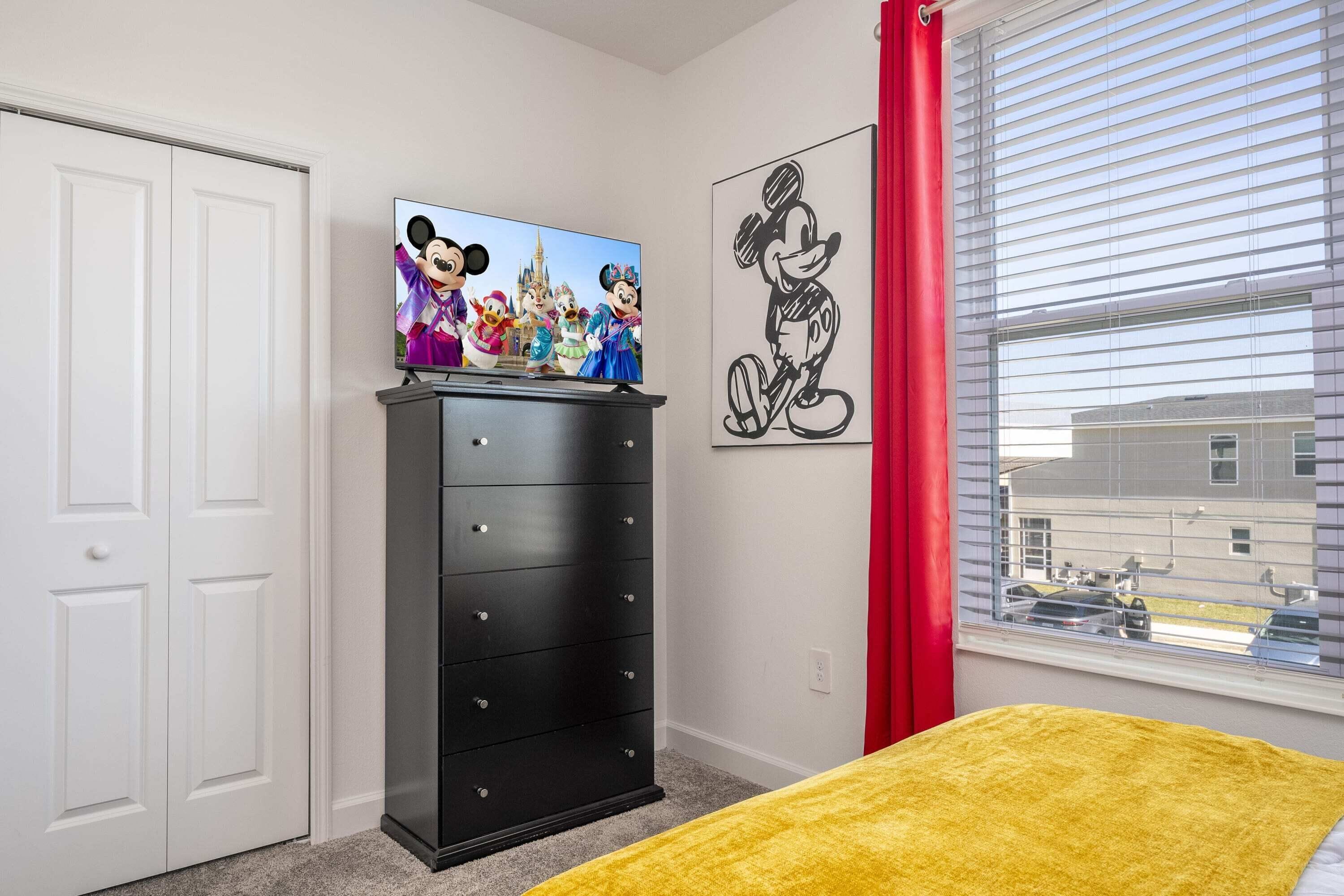 A bedroom with a TV on a dresser, a Mickey Mouse poster, and a window with blinds.