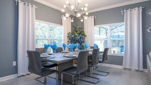 Uma sala de jantar com uma mesa grande, seis cadeiras, um candelabro e vista para o exterior.