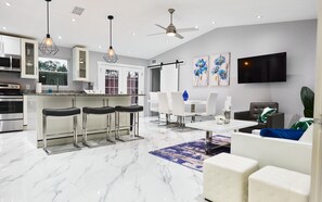 A modern kitchen with bar stools, a dining area with a TV, and a living room with a sofa and a coffee table.