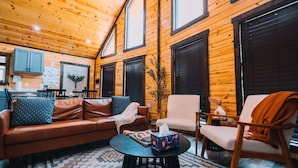 Un interior de cabaña acogedor, con paredes de madera, una sala de estar con un sofá de cuero marrón, un sillón blanco y una mesa de centro redonda.