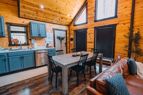 A cozy cabin interior with a wooden interior, a dining area with a table and chairs, a kitchen with blue cabinets, and a leather couch.