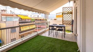 En balkon med kunstgræs, et bord med stole og udsigt til omkringliggende bygninger.