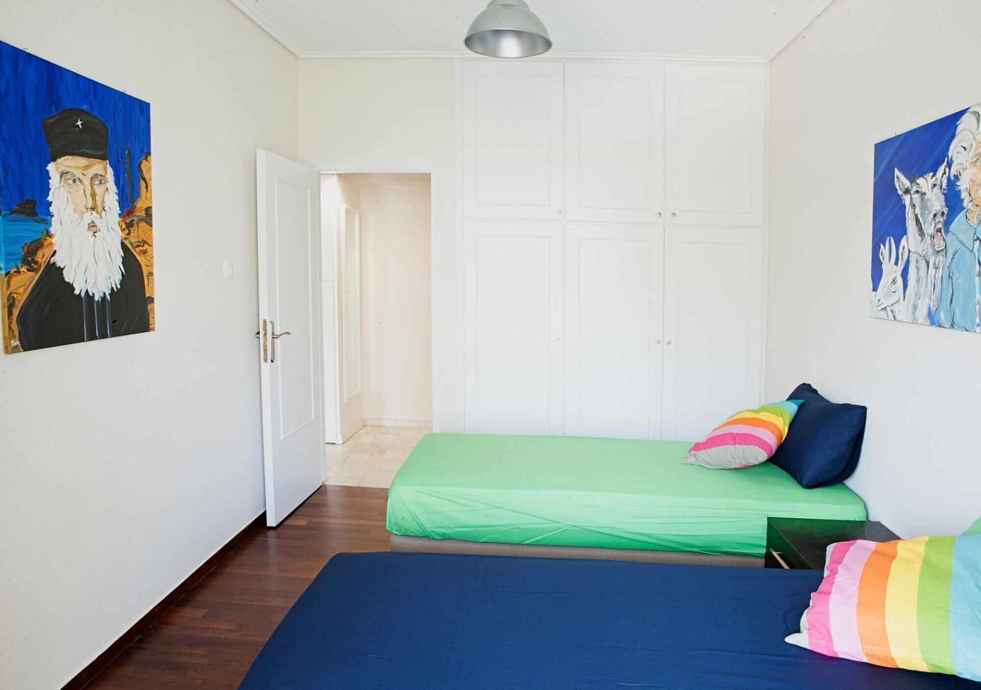 Een kamer met een bed, een groene matras, een blauwe matras en kleurrijke kussens. Er is een witte deur en een witte garderobe.
