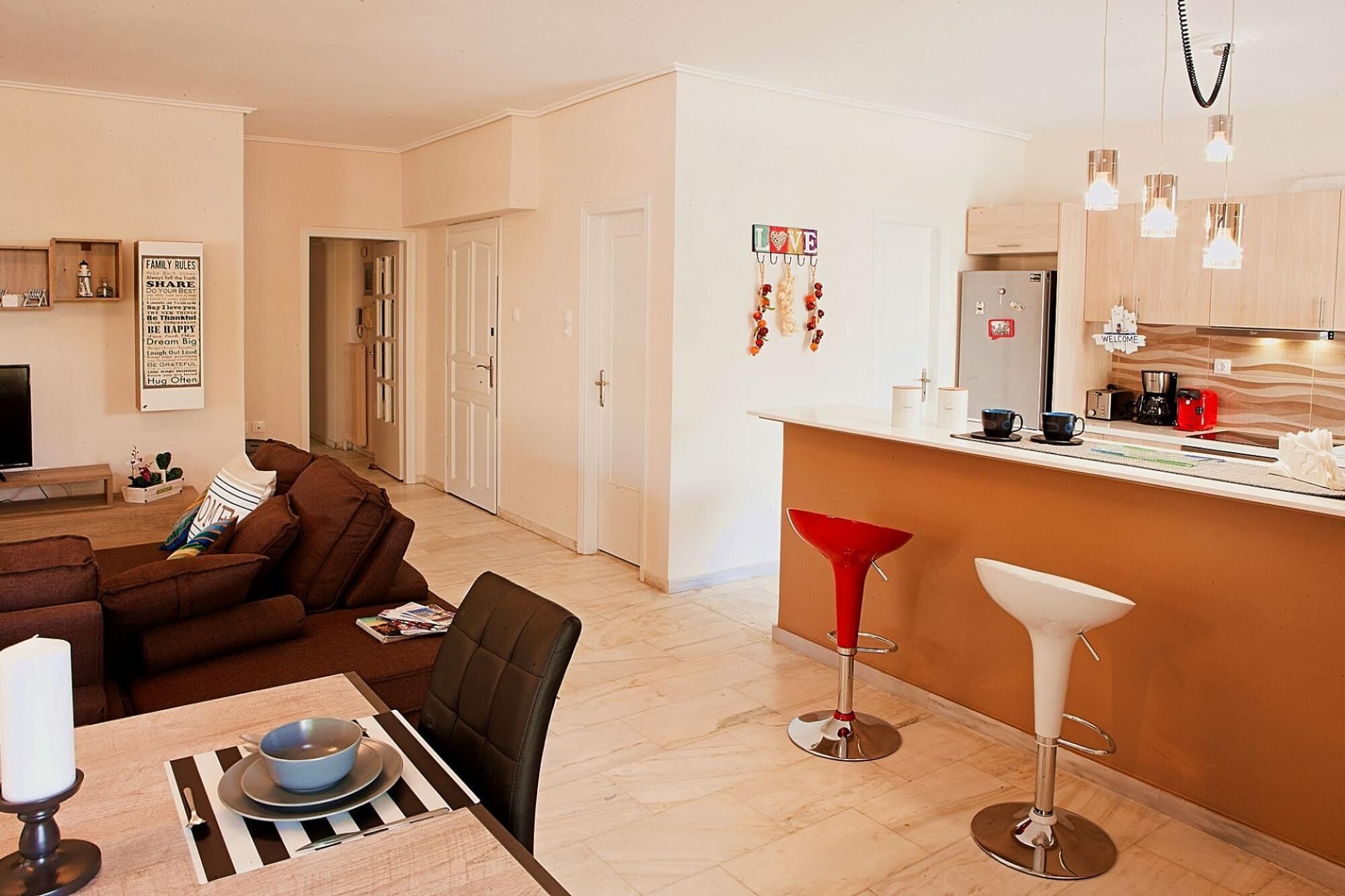 A modern living room with a brown sofa, a dining table with a bowl and cutlery, and a kitchen area with a bar stool and pendant lights.