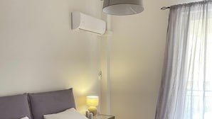 A hotel room with two beds, a bedside table with a lamp, an air conditioning unit, and a window with curtains. - Cassiopeia - Glyfada Apartment (Glyfada)