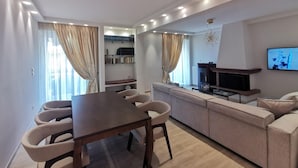 A modern living room with a dining area, a sofa set, a fireplace, and a TV mounted on the wall. - Cassiopeia - Glyfada Apartment (Glyfada)