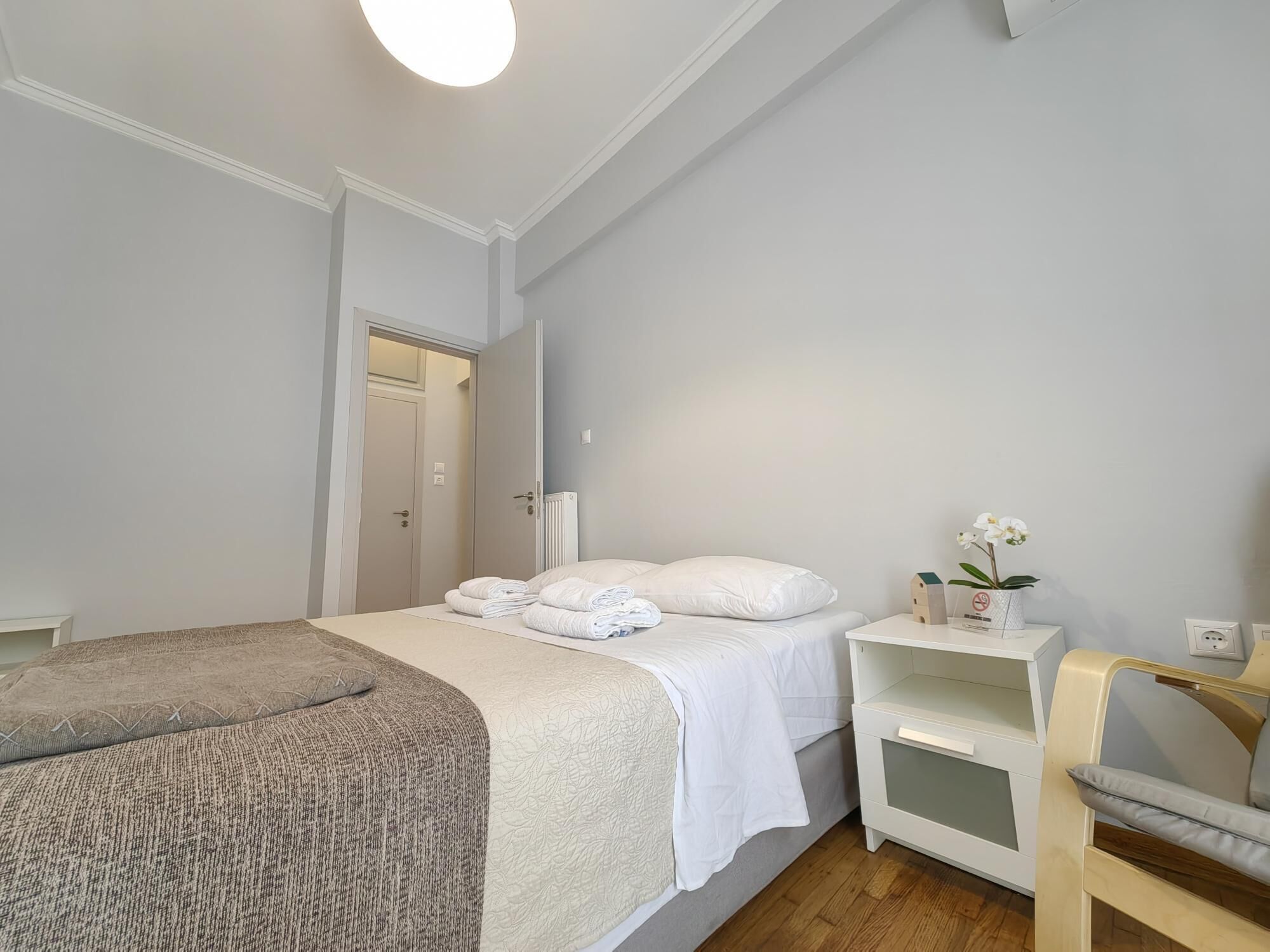 Ein Schlafzimmer mit einem Bett, einem Nachttisch mit Lampe, einem Sessel und einem kleinen Regal mit einer Orchidee.