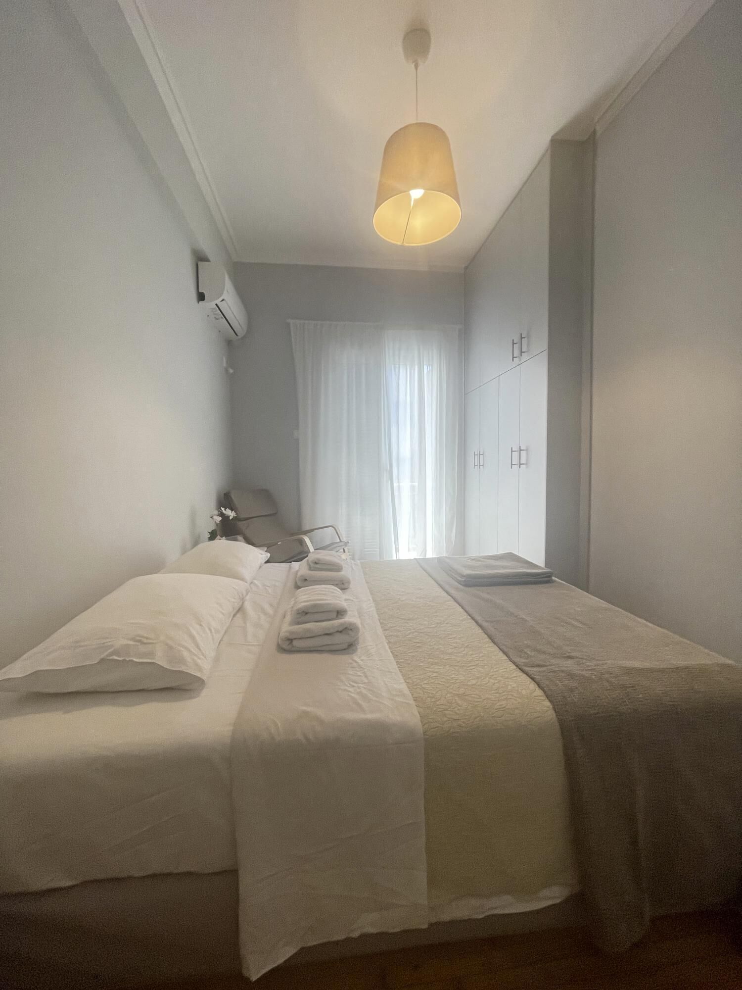 A hotel room with two single beds, a bedside table with a lamp, a window with curtains, and a hanging light fixture.