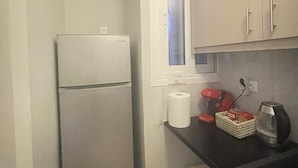 Una cocina compacta con refrigerador, una ventana con persianas, un rollo de papel higiénico, una tetera roja, una cafetera y una cesta de pan.