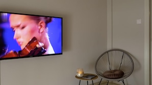 A modern living room with a flat-screen TV mounted on the wall, a round chair with a cushion, a small table with a candle and a book, and a pendant light fixture. - Demetra - Koukaki Center Apartment (Athens)