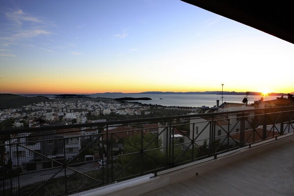 A balcony with a view of a coastal town at sunset. - Libra - Luxurious Sea View Apartment (Vari-Voula-Vouliagmeni)