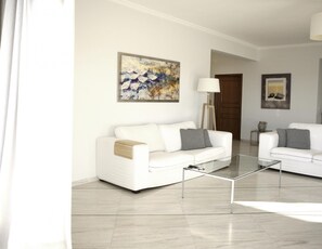A modern living room with a white sofa, a glass coffee table, and a painting on the wall. - Libra - Luxurious Sea View Apartment (Vari-Voula-Vouliagmeni)
