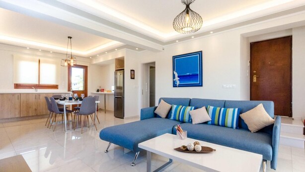 A modern living room with a blue sofa, a white coffee table, and a dining area with a kitchen in the background. - Leo - Unique Glyfada Apartment (Glyfada)