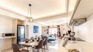 A modern kitchen with a refrigerator, microwave, and coffee maker. There is a dining area with a table set for two and a sofa. - Leo - Unique Glyfada Apartment (Glyfada)