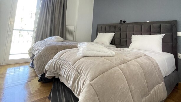 A bedroom with two beds, a grey headboard, a hanging light fixture, and a window with curtains.