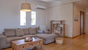 Un salon moderne avec un canapé beige, une table basse en bois et un luminaire suspendu. On y trouve également une bibliothèque et un radiateur fixés au mur.