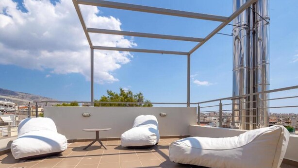 A rooftop terrace with white cushioned seating, a small table, and a view of the city and mountains. - Solon - Deluxe City Center Apartment (Agios Dimitrios)