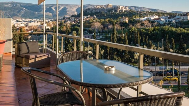 A balcony with wicker furniture, a glass table, and a view of a cityscape with mountains in the background. - Virgo - Loft With Spectacular View to Acropolis (Athens)