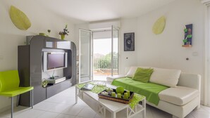 Un salon moderne avec un canapé blanc, un fauteuil vert, une table basse sur laquelle se trouve un plateau avec divers objets, un téléviseur posé sur un meuble et un balcon doté d’une rambarde.