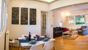 A modern living room with a dining area, a sofa, and a coffee table. There are three paintings on the wall, a window with a view of greenery, and a door leading to another room. - Aquarius - Glyfada Detached House (Glyfada)