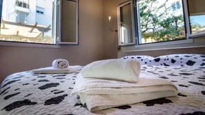 A bedroom with a bed, two windows, and a view of buildings and trees outside. - Aquarius - Glyfada Detached House (Glyfada)