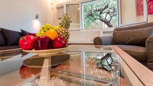 A modern living room with a glass coffee table, a fruit bowl, a brown leather sofa, and a large window with a tree view. - Aquarius - Glyfada Detached House (Glyfada)