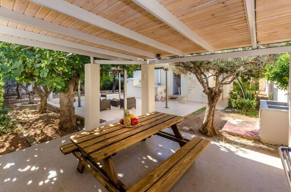 An outdoor patio with a wooden table and benches, surrounded by trees and a covered area with a view of a building in the background.