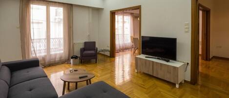 A spacious living room with a large sofa, a TV on a wooden stand, and a balcony with curtains.