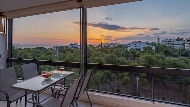 A balcony with a table and chairs, overlooking a cityscape with buildings and trees during sunset. - Archer - Sea View Apartment (Vari-Voula-Vouliagmeni)