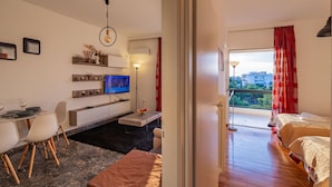 A modern hotel room with a dining area, a TV, a balcony with red curtains, and a view of the city. - Archer - Sea View Apartment (Vari-Voula-Vouliagmeni)