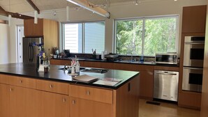 A modern kitchen with wooden cabinets, stainless steel appliances, a large island with a sink, and a view of trees through the windows. - Hidden Gem on the prairie, a house of light and ease (Washington)