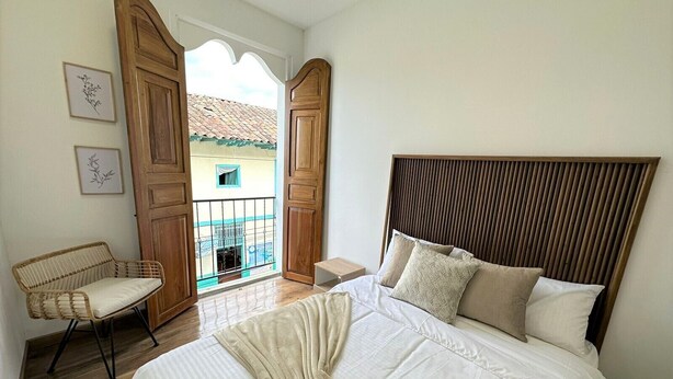 A bedroom with a large bed, a wooden headboard, a wicker chair, and a balcony with a view of buildings. - Casa Hotel Real (Circasia)