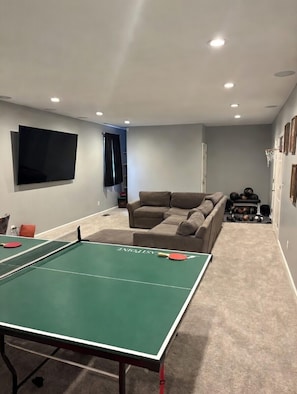 A living room with a ping pong table, a large flat-screen TV, a couch, and a basketball hoop.