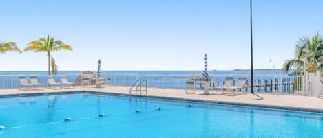 A swimming pool with clear blue water, surrounded by lounge chairs and palm trees, with a view of the ocean.