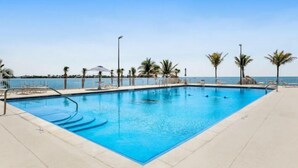 Piscina retangular com água azul cristalina, rodeada por um deck revestido e palmeiras.