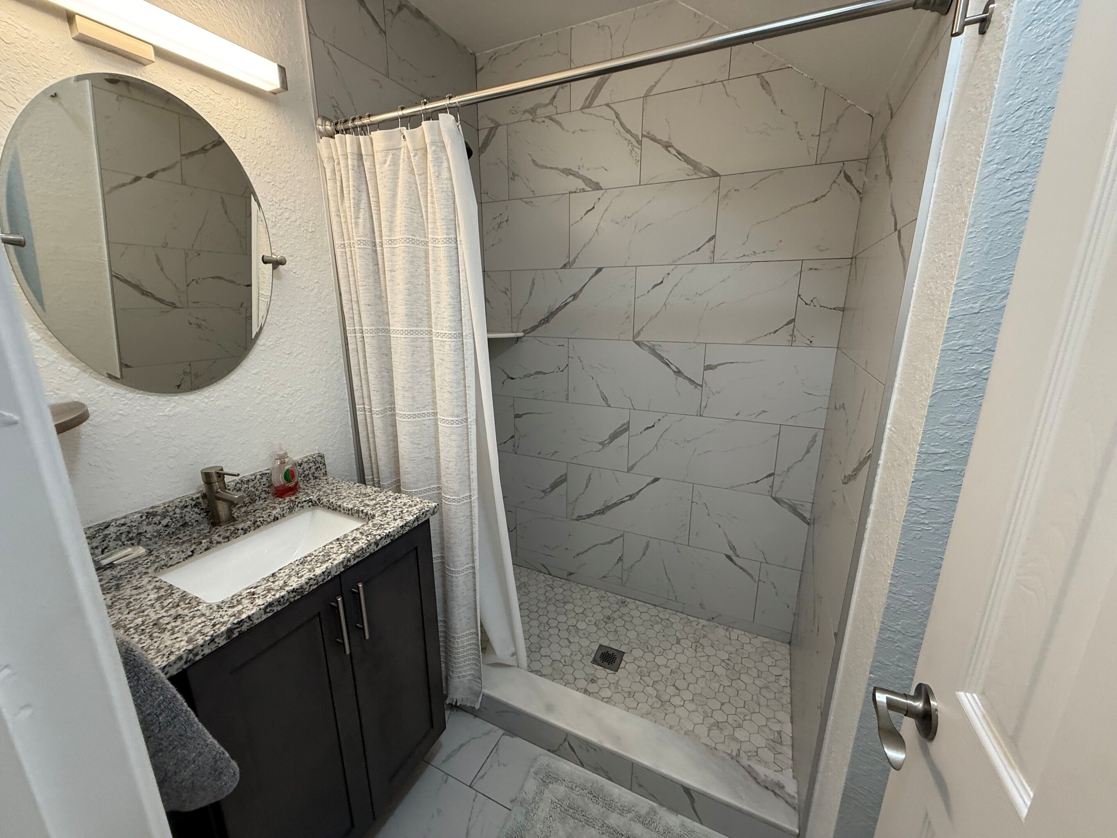A bathroom with a granite countertop, a sink, a mirror, and a shower with a marble wall.