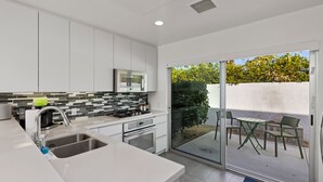 A modern kitchen with a large sink, stove, and oven, a tiled backsplash, and a sliding glass door leading to an outdoor patio with chairs. - Come visit Palm Springs! (Palm Springs)