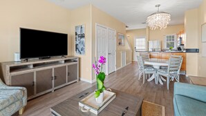 A living room with a flat-screen TV, a dining table with chairs, a coffee table, and a decorative orchid plant.