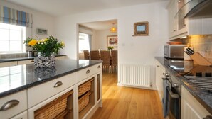 A modern kitchen with white cabinets, a granite countertop, and a built-in oven. There is a dining area with wicker chairs and a table, and a small plant on the countertop. - Ty Coed (Pwllheli)