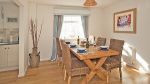 A dining area with a wooden table and wicker chairs, a vase with branches, and a window with curtains. - Ty Coed (Pwllheli)