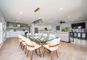 A modern kitchen with white cabinets, a dining area with a glass table and white chairs, and a ceiling fan.