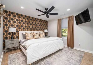 A bedroom with a bed, a ceiling fan, a TV mounted on the wall, and a window with curtains.