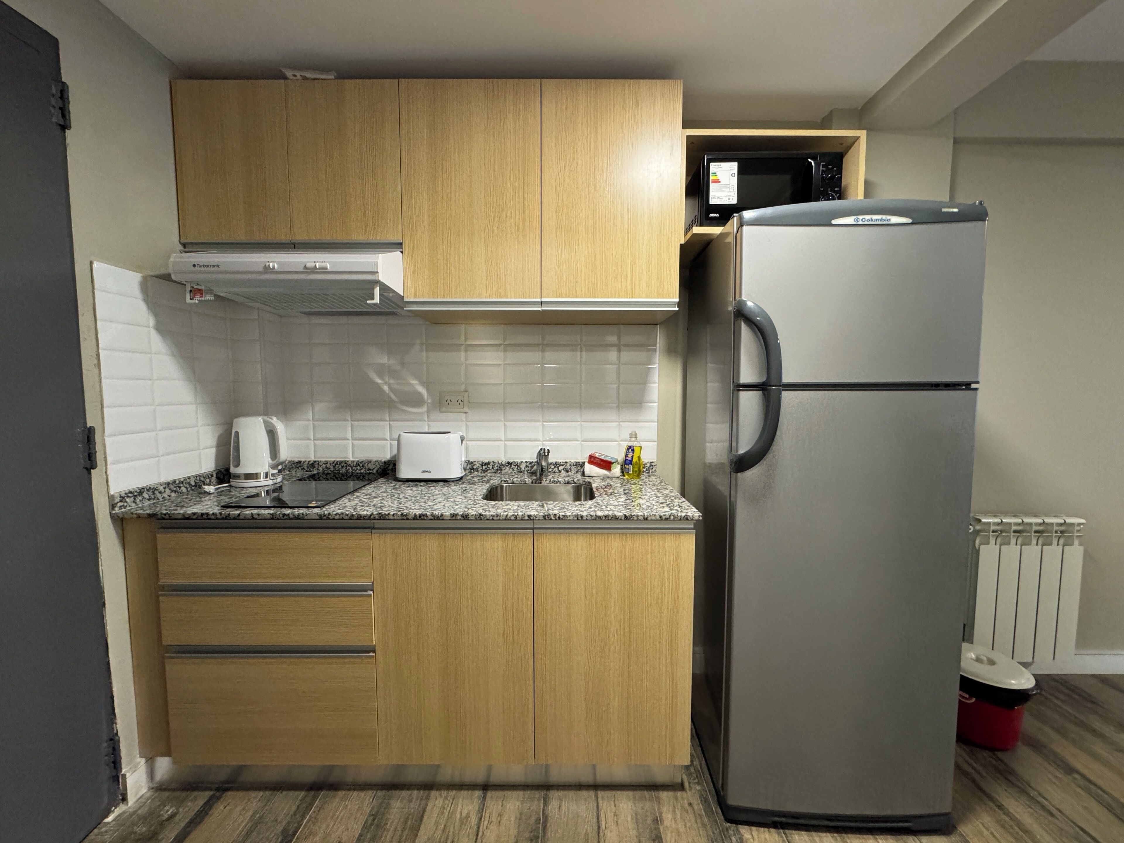 A compact kitchen with wooden cabinets, a stainless steel refrigerator, a microwave, a toaster, and a kettle.