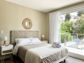 A neatly made bed with a headboard, two bedside tables with lamps, a round mirror, and a view of greenery through a large window.