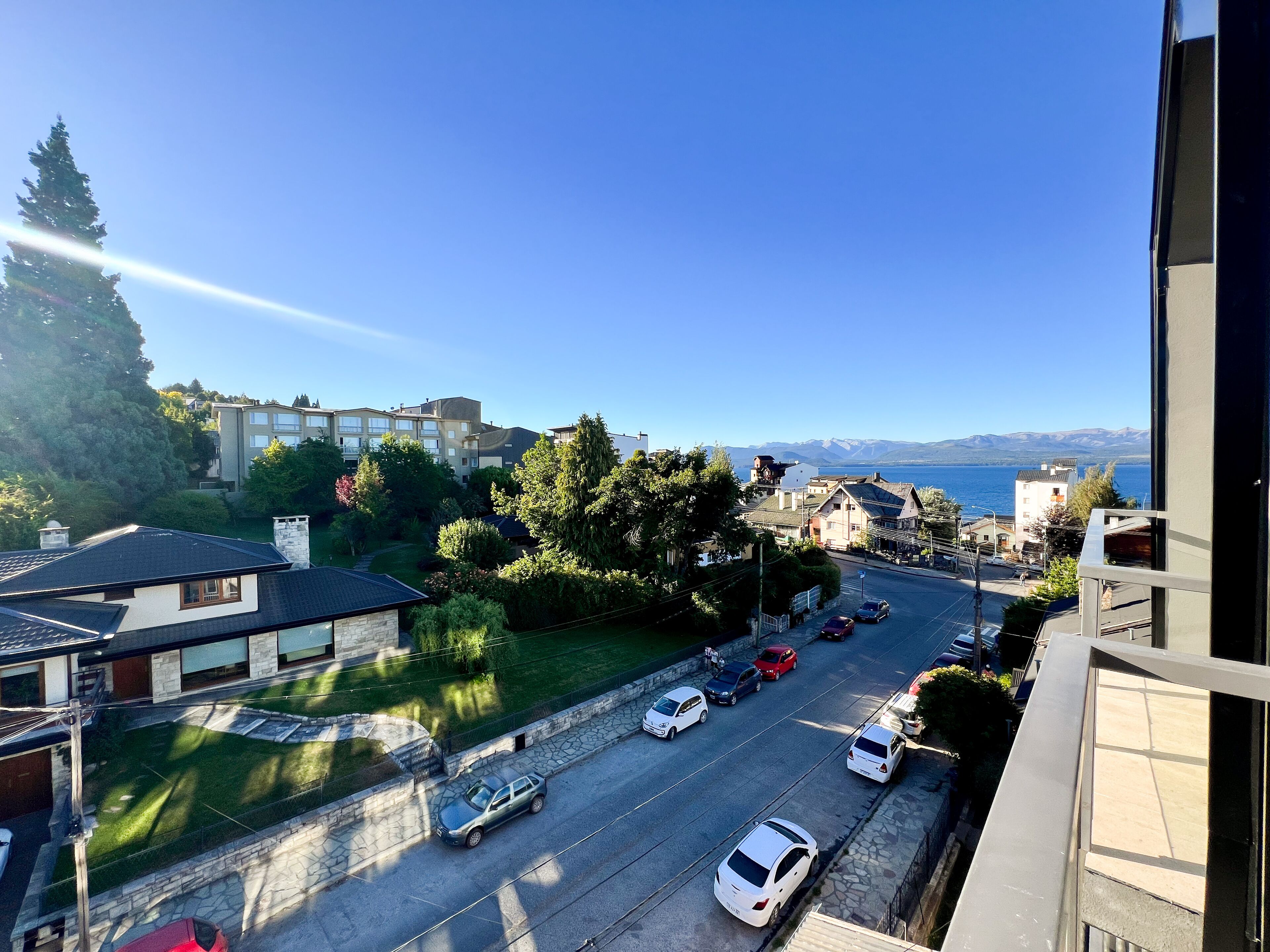 Une rue résidentielle bordée de maisons, un ciel dégagé et une vue lointaine sur des montagnes et de l’eau.