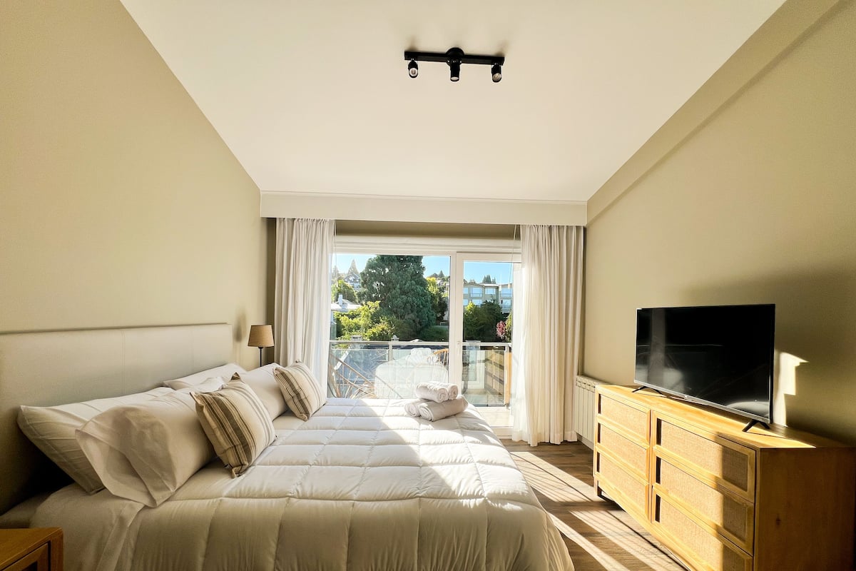 Ein Hotelzimmer mit einem großen Bett, einem Fernseher, einer hölzernen Kommode und einem Balkon mit Blick auf die Stadt.