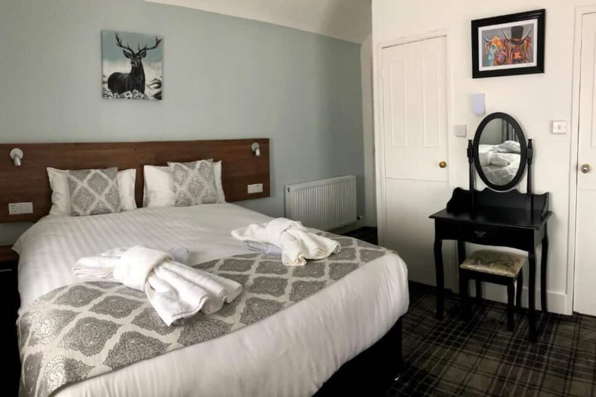 A neatly made bed with patterned bedding, a wooden headboard, a bedside table with a mirror, and a framed picture on the wall.